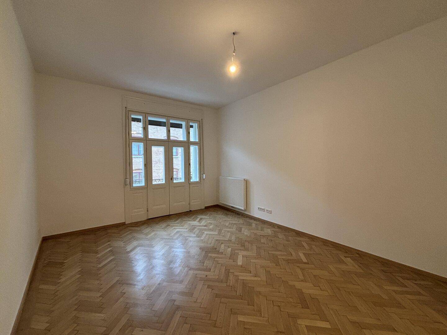 Großer, heller Raum mit Fischgrätparkett und Balkonzugang, ideal als Schlaf- oder Wohnzimmer.