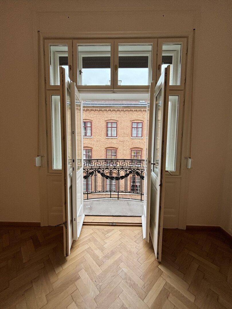Blick durch die geöffneten Balkontüren auf einen kleinen Balkon mit schmiedeeisernem Geländer und gegenüberliegendem Gebäude.