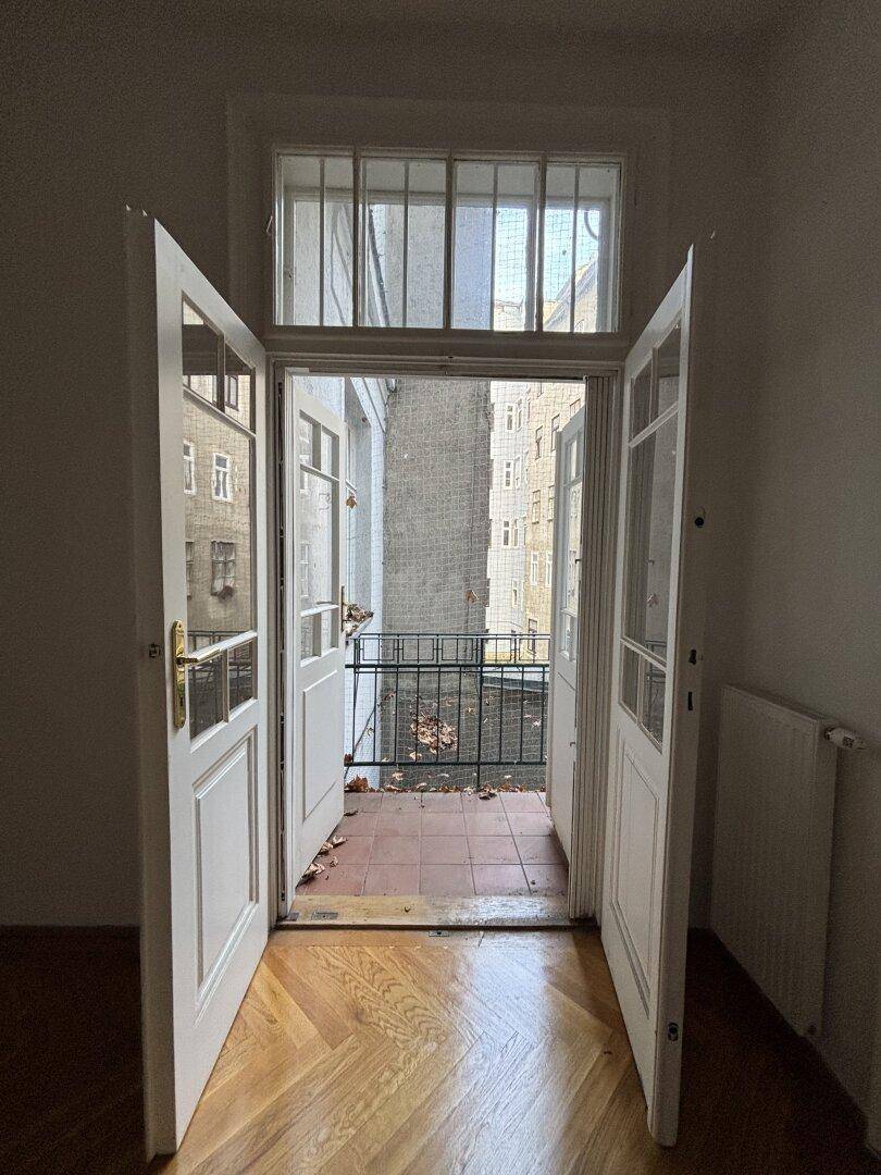 Blick aus einem Zimmer durch offene Doppeltüren auf einen kleinen Balkon mit Terrakottafliesen und Stadtblick.