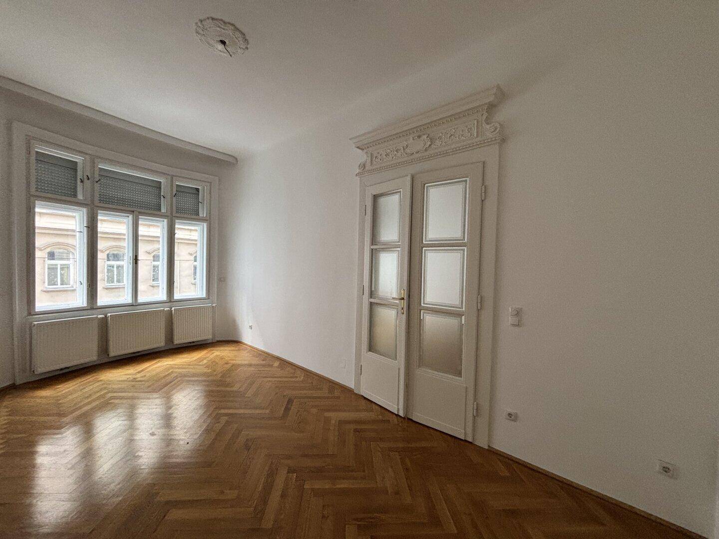 Helles Zimmer mit Fischgrätparkett und einer eleganten Doppeltür mit Zierrahmen, ideal für Wohn- oder Schlafbereich.