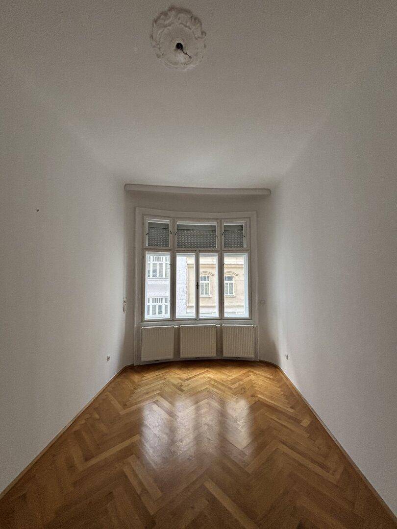Gemütliches Zimmer mit klassischem Fischgrätparkett, Stuckdecke und großem Fenster, das viel Licht hereinlässt.