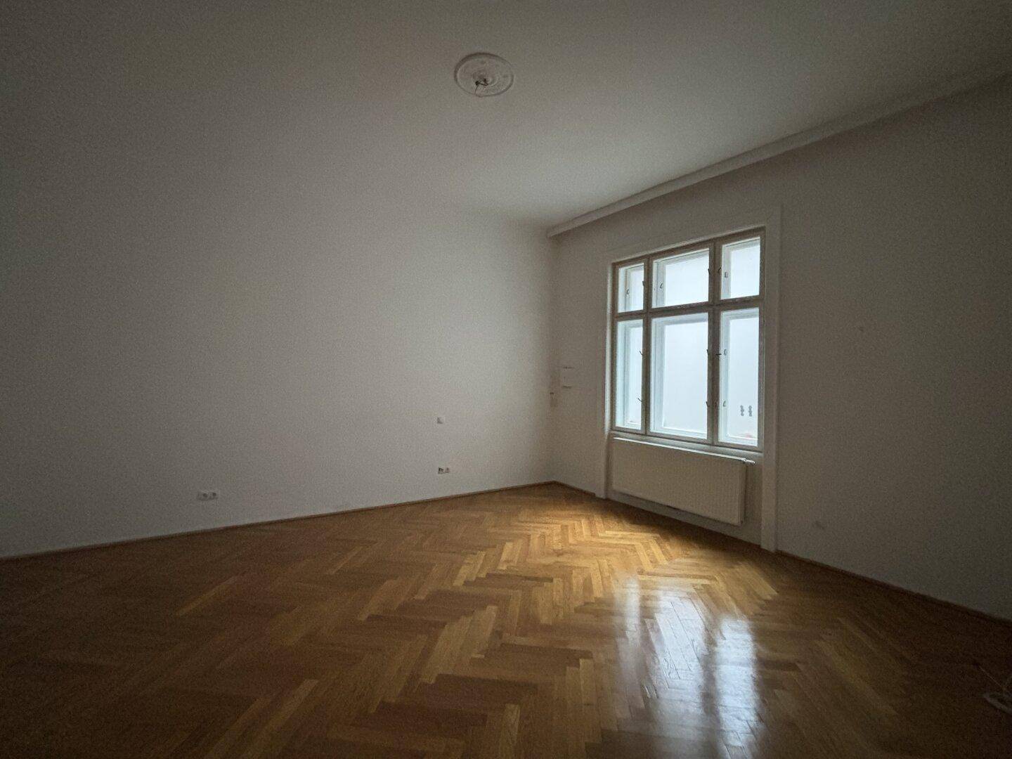 Helles Schlafzimmer mit klassischem Fischgrätparkett, Stuckdecke und großem Fenster, das viel Tageslicht bietet.