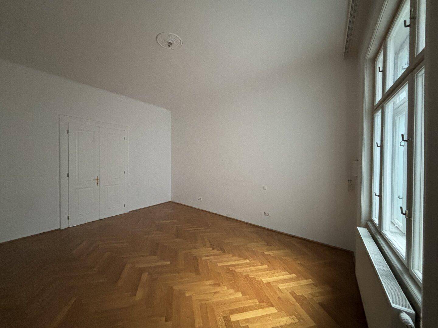 Geräumiges Zimmer mit Fischgrätparkett und großen Fenstern, die für eine helle und freundliche Atmosphäre sorgen.
