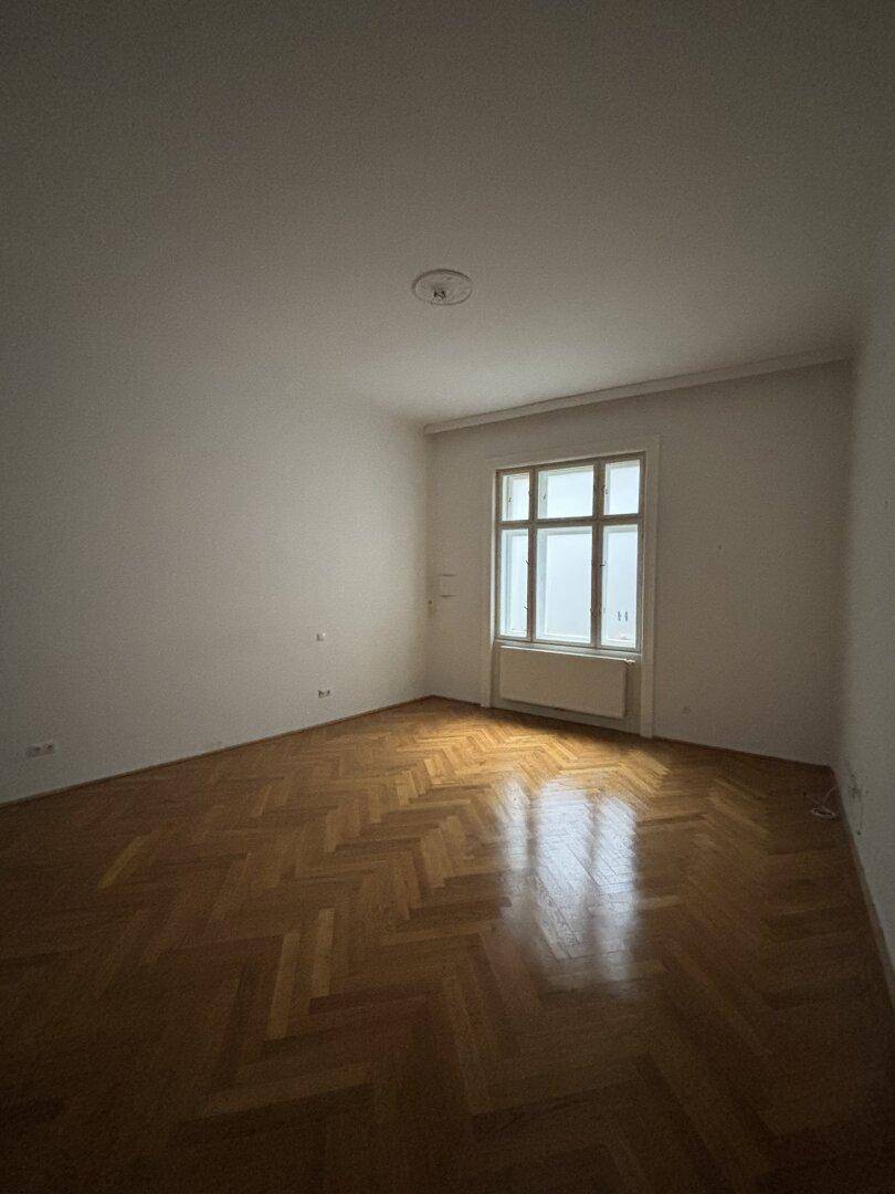 Ruhiges Zimmer mit klassischem Fischgrätparkett, Stuckdecke und Fenster, ideal als Schlaf- oder Arbeitszimmer.