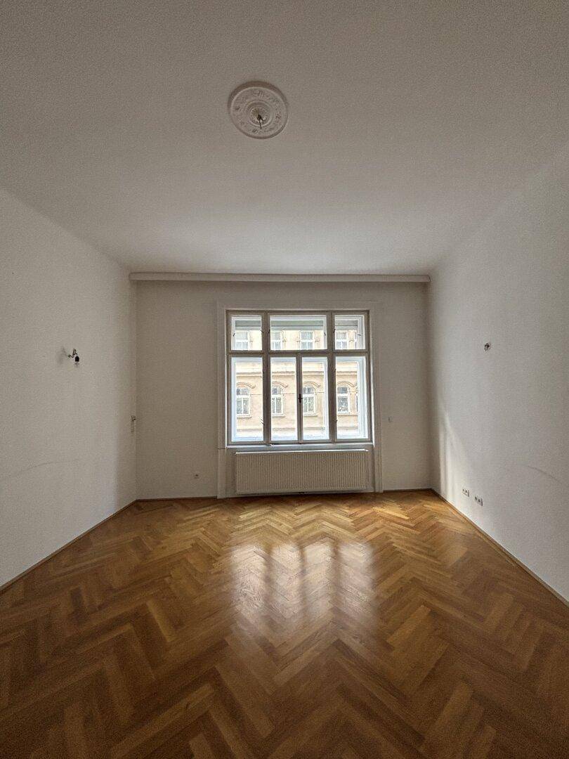 Helles Zimmer mit Fischgrätparkett, Stuckdecke und großem Fenster, das einen Blick auf die Stadt bietet.