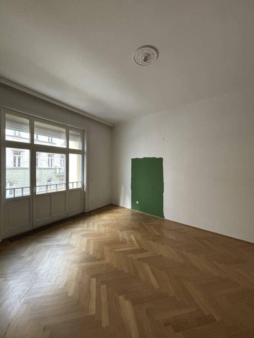 Geräumiges Zimmer mit Fischgrätparkett, grüner Akzentwand und Zugang zu einem kleinen Balkon.