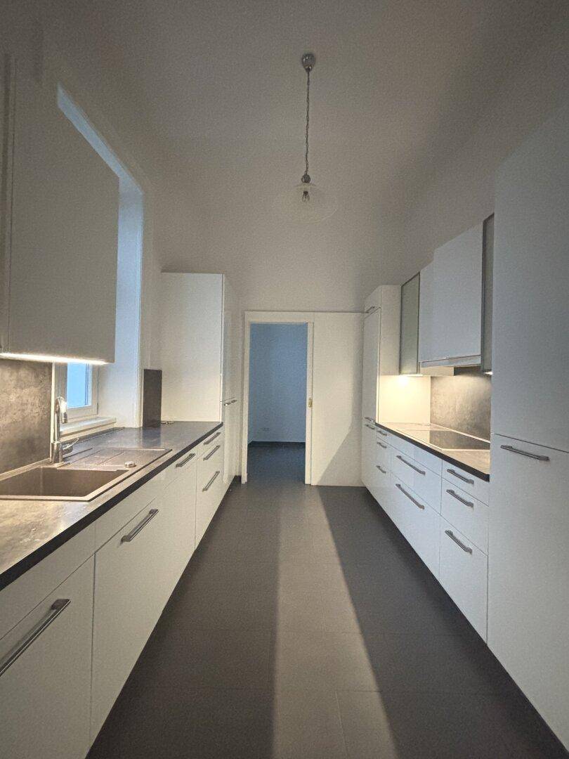 Moderne, schmale Küche mit weißen Schränken, dunklen Arbeitsplatten und grauen Fliesen, gut beleuchtet.