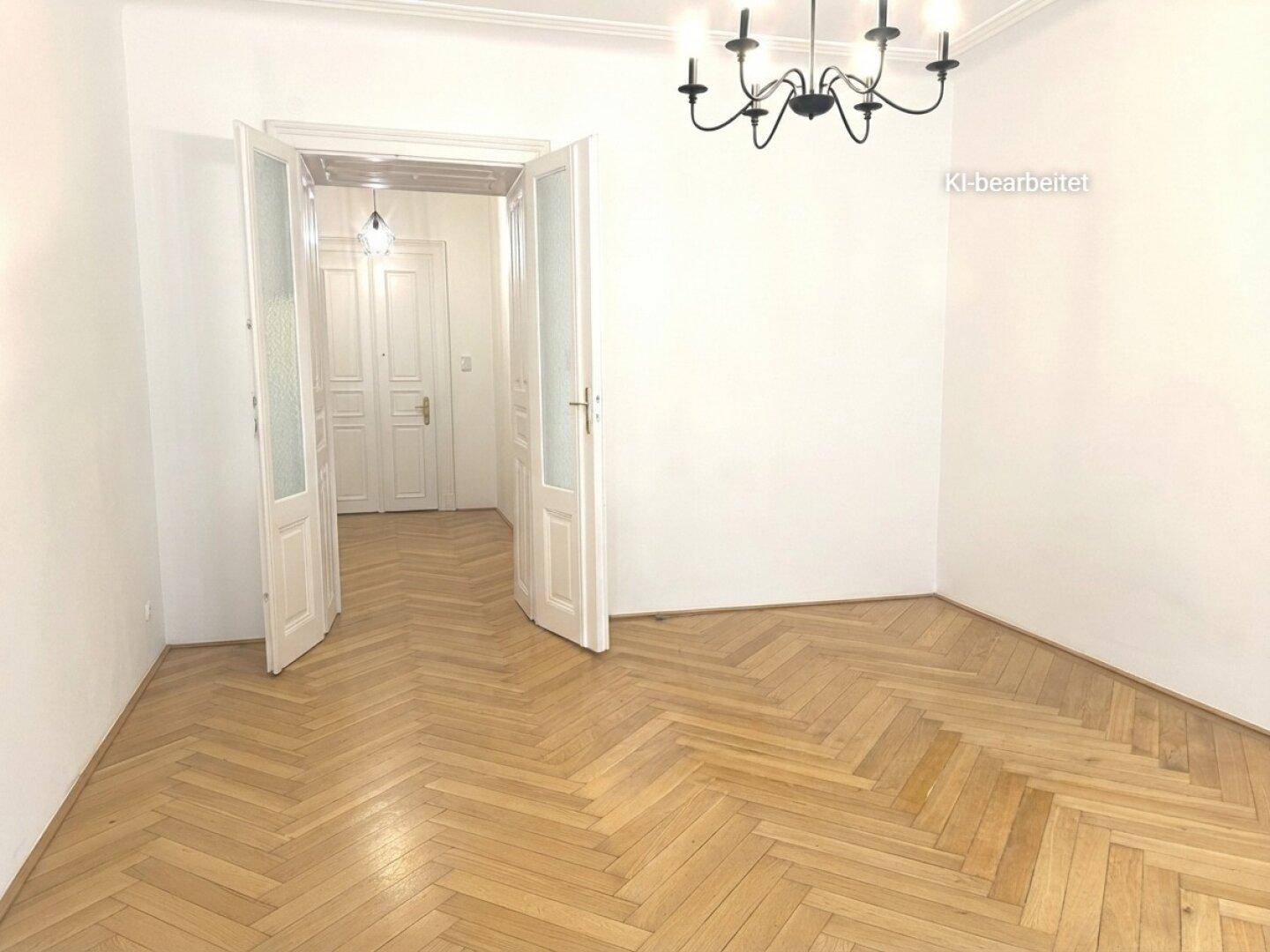 Geräumiges Zimmer mit Fischgrätparkett, hohen Decken und doppelten Flügeltüren zu einem weiteren Raum.