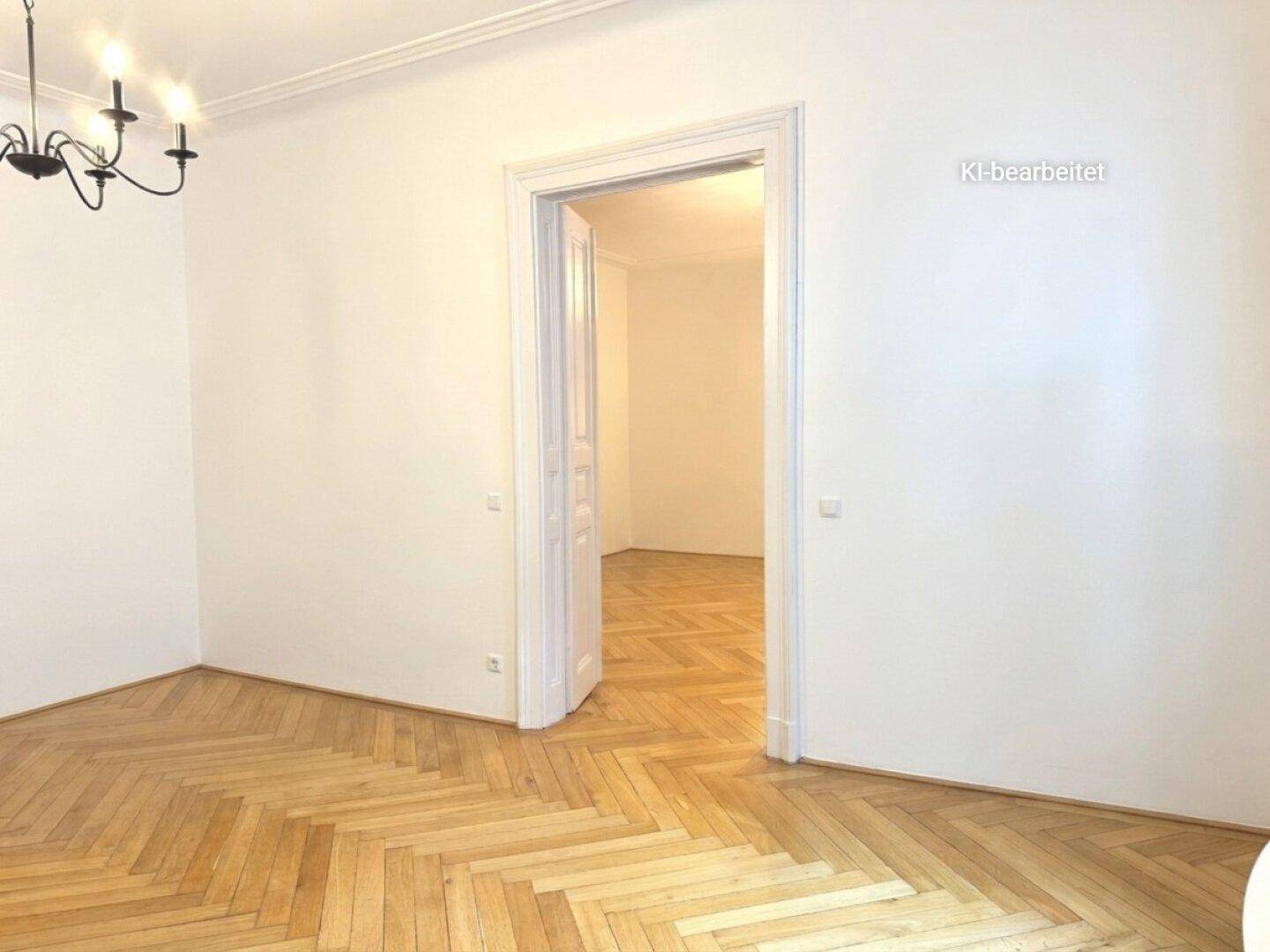 Heller Raum mit Fischgrätparkett, hohen Decken und einer eleganten Türöffnung zu einem Nebenraum.
