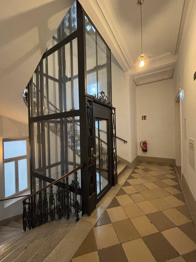 Historisches Treppenhaus mit modernem Glasaufzug, hohen Decken und gemustertem Fliesenboden.