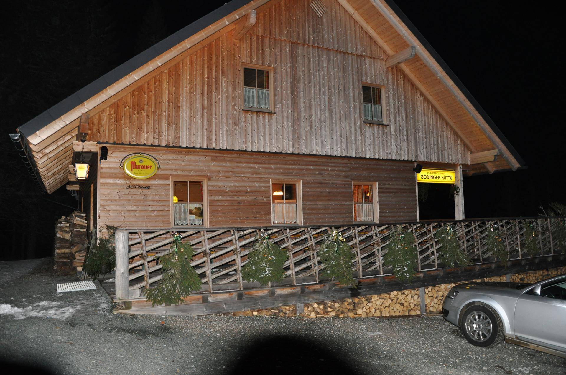 Holzhütte bei Nacht mit beleuchteten Fenstern und einem geparkten Auto, umgeben von dunkler Landschaft.