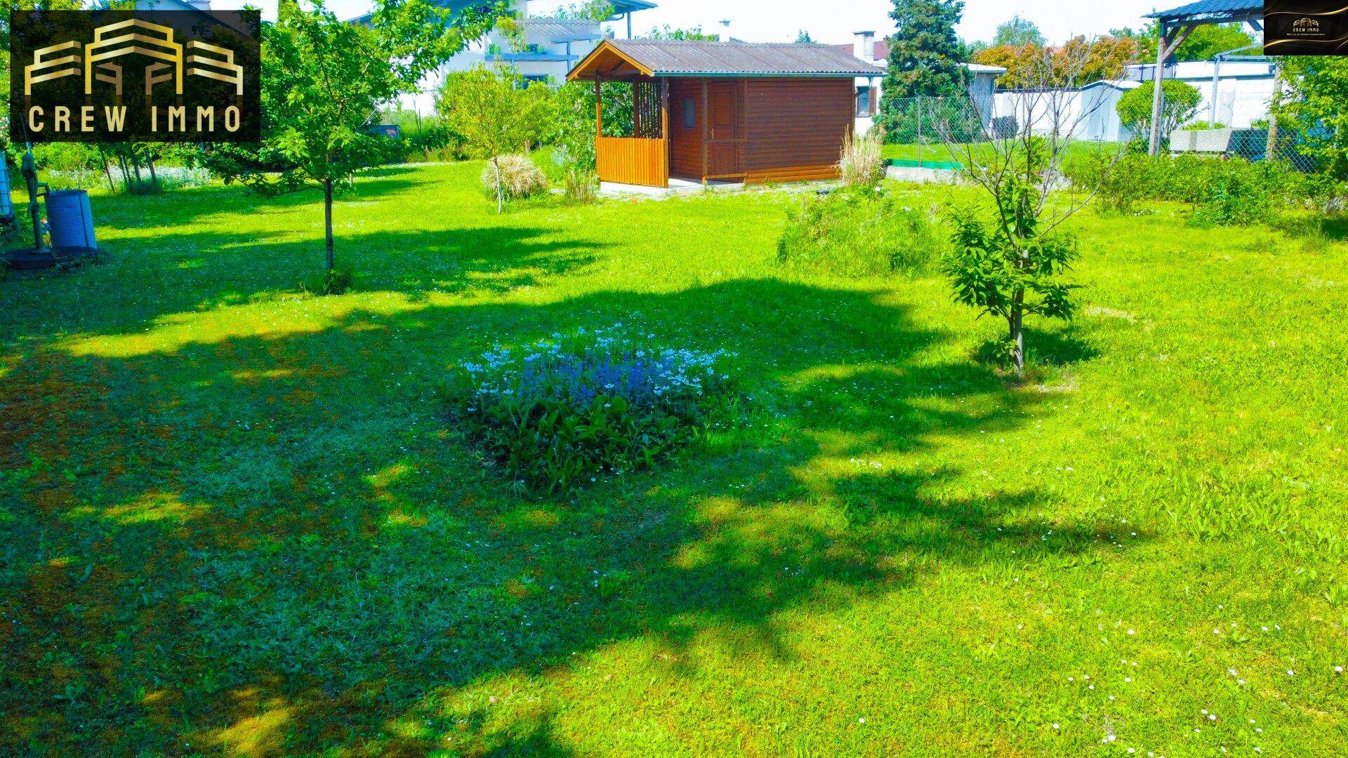 Weitläufige Rasenfläche mit blühenden Pflanzen und einem charmanten Gartenhaus im Sonnenlicht.