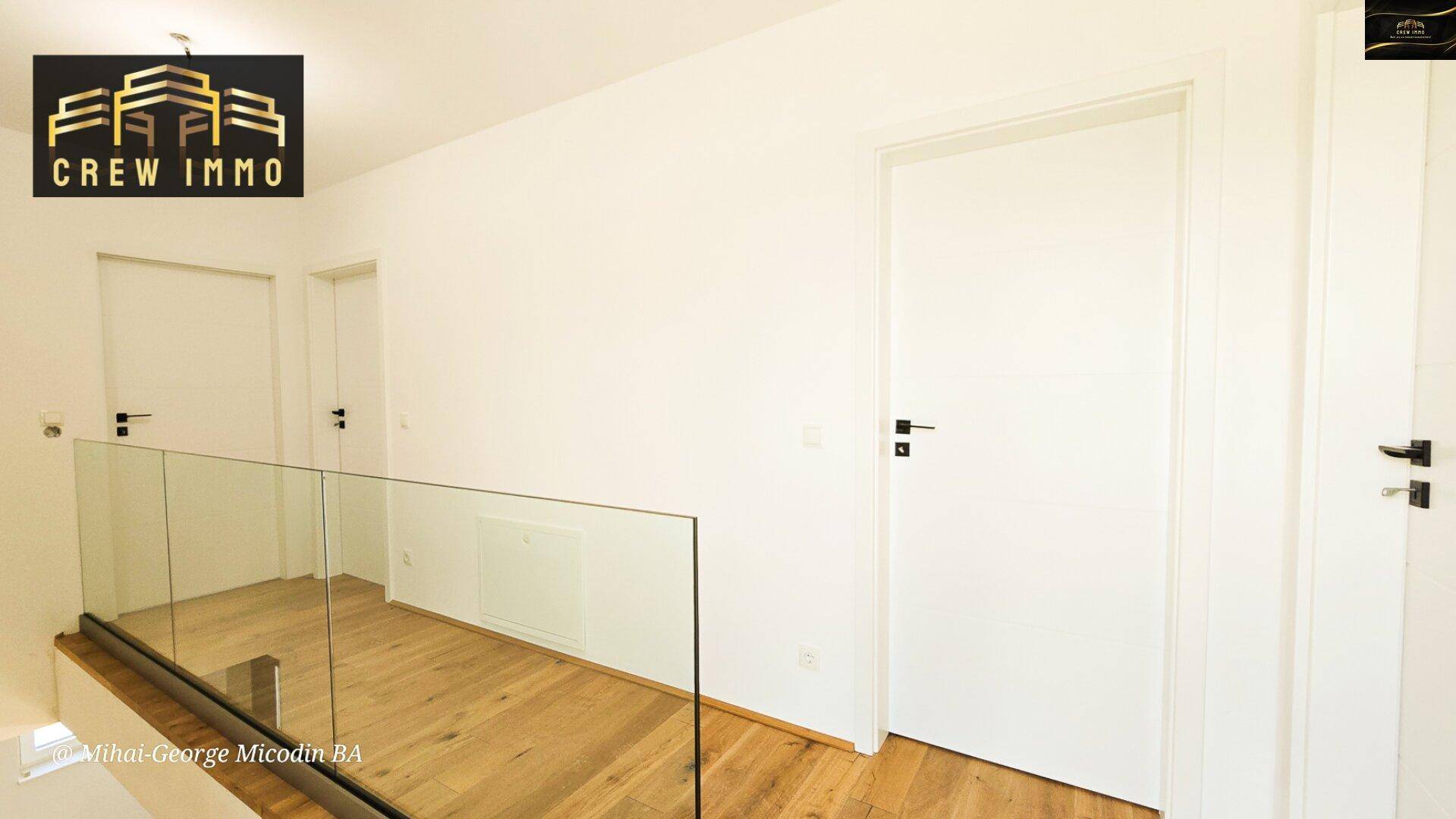 Heller Flur im Obergeschoss mit Holzboden, Glasgeländer und weißen Türen, die zu den Zimmern führen.