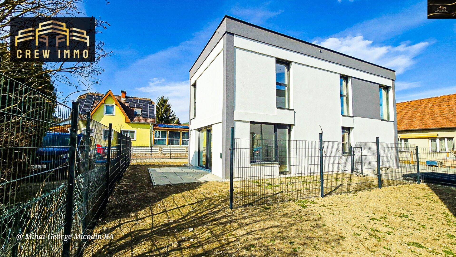 Rückseite des modernen Hauses mit kleiner Terrasse und angrenzendem Gartenbereich, umgeben von einem Zaun.