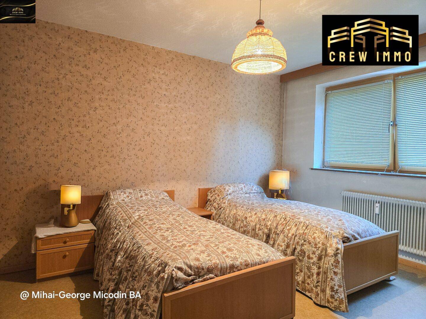 Schlafzimmer mit zwei Einzelbetten, gemusterter Tapete und Fenster mit Jalousien.