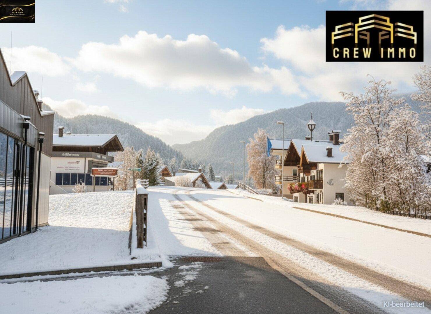 Verschneite Straße in einer alpinen Siedlung mit Häusern und Bergen im winterlichen Hintergrund.
