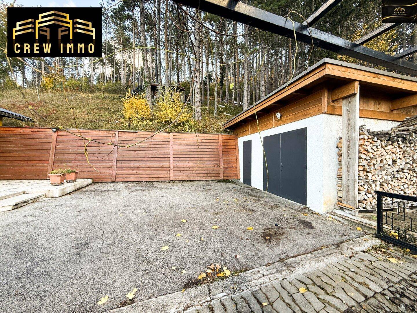 Garage mit Holzverkleidung und großem Vorplatz, umgeben von Bäumen und Natur.