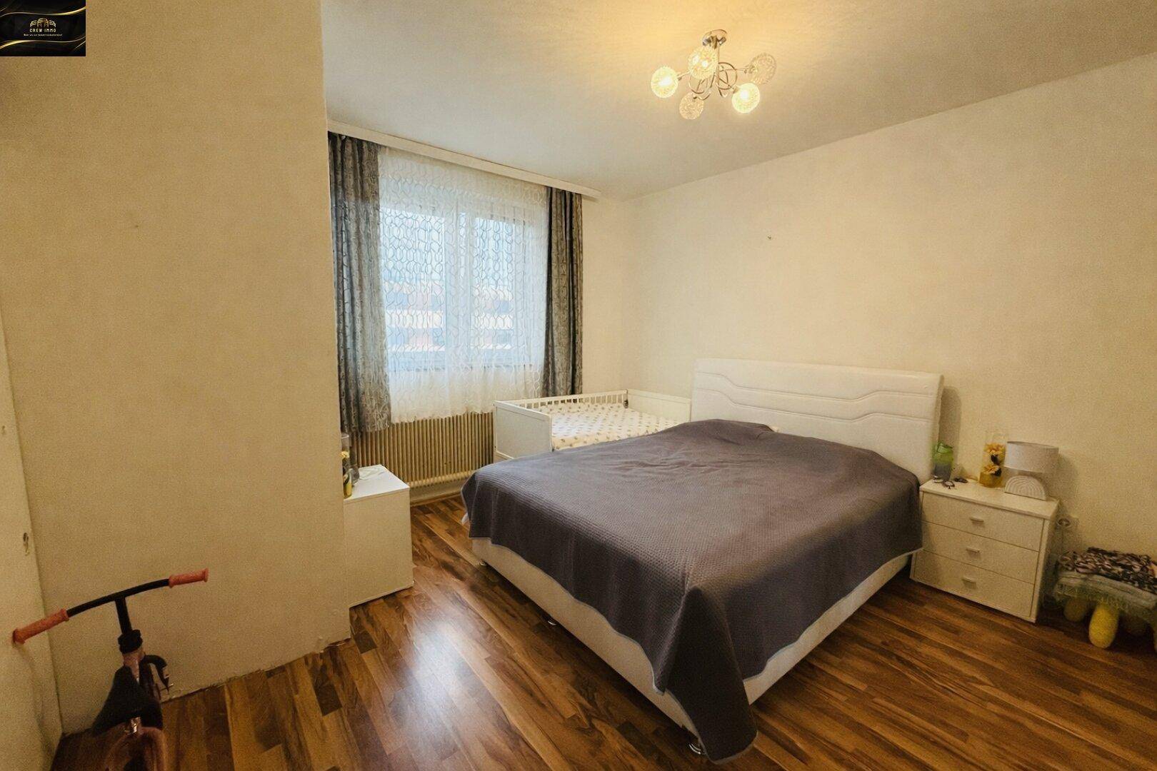 Schlafzimmer mit Doppelbett, Babybett und Laminatboden, beleuchtet durch ein Fenster.