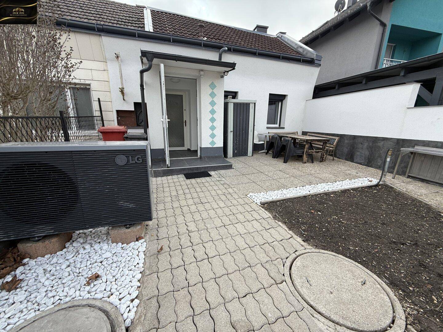 Gepflasterte Terrasse mit kleiner Gartenfläche und einer Wärmepumpe im Außenbereich.