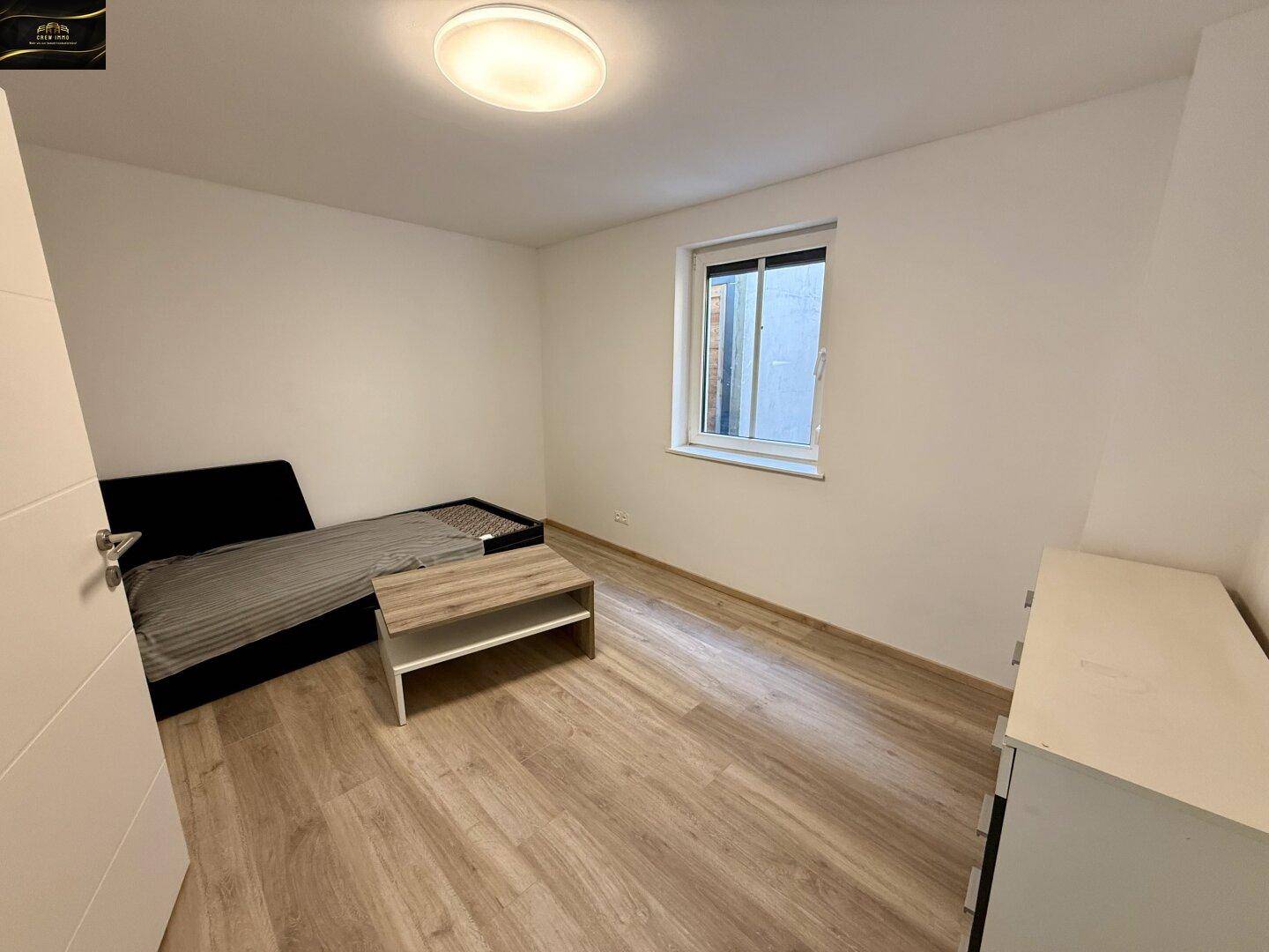 Helles Schlafzimmer mit Laminatboden, einem einfachen Bett und einem Fenster.