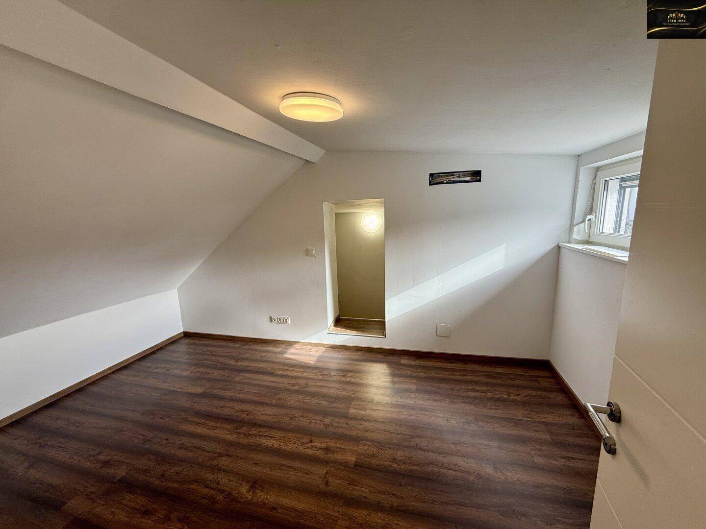 Helles Zimmer im Dachgeschoss mit schrägen Wänden, Laminatboden und Fenster.