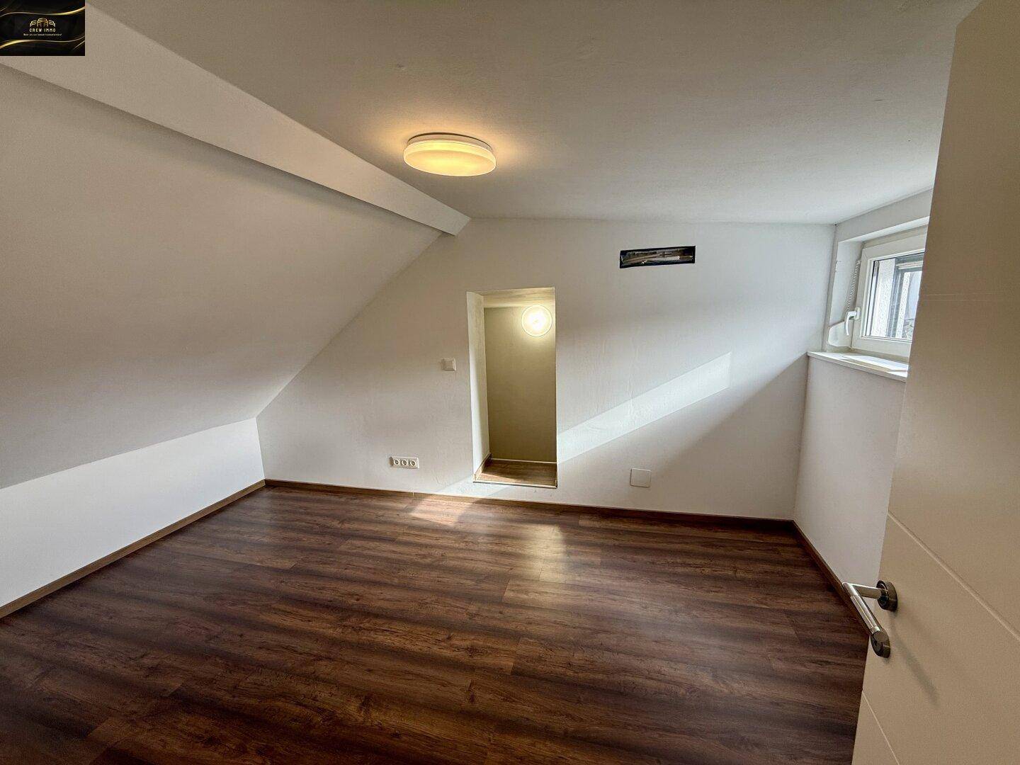 Gemütliches Zimmer im Dachgeschoss mit schrägen Wänden, Laminatboden und Fenster.