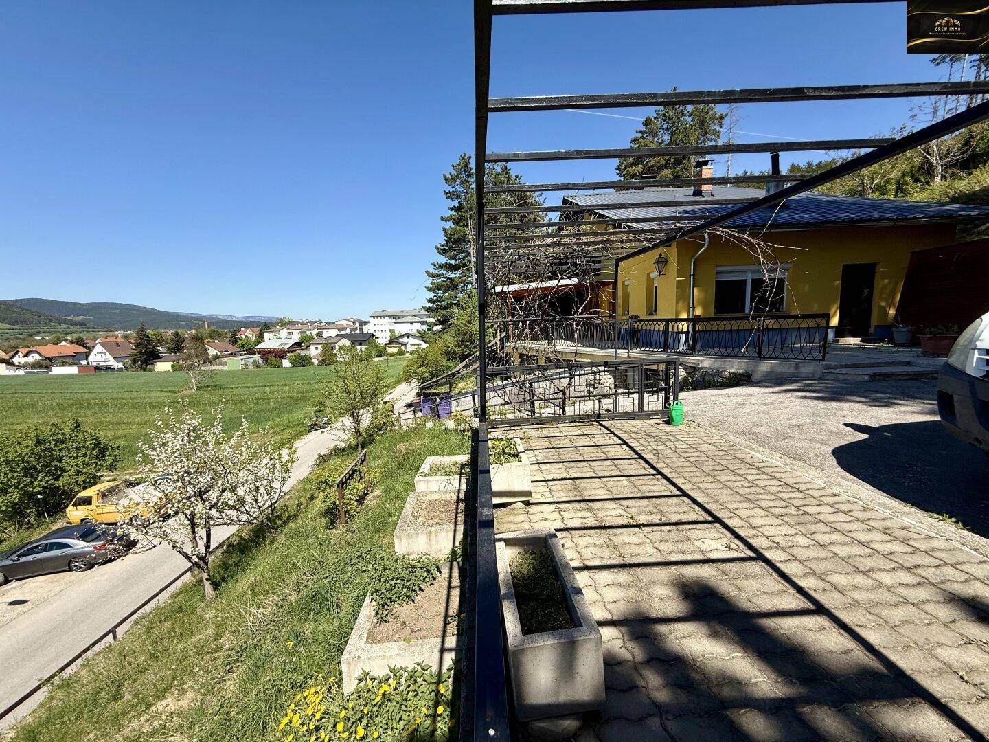 Gepflasterte Terrasse mit Pergola-Struktur und weitem Blick über die Landschaft und umliegende Häuser.