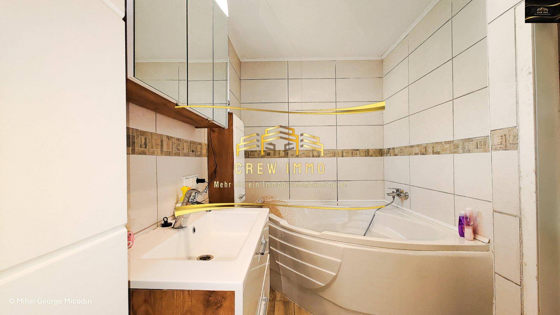Badezimmer mit Eckbadewanne, Waschbecken und Spiegelschrank, komplett gefliest.