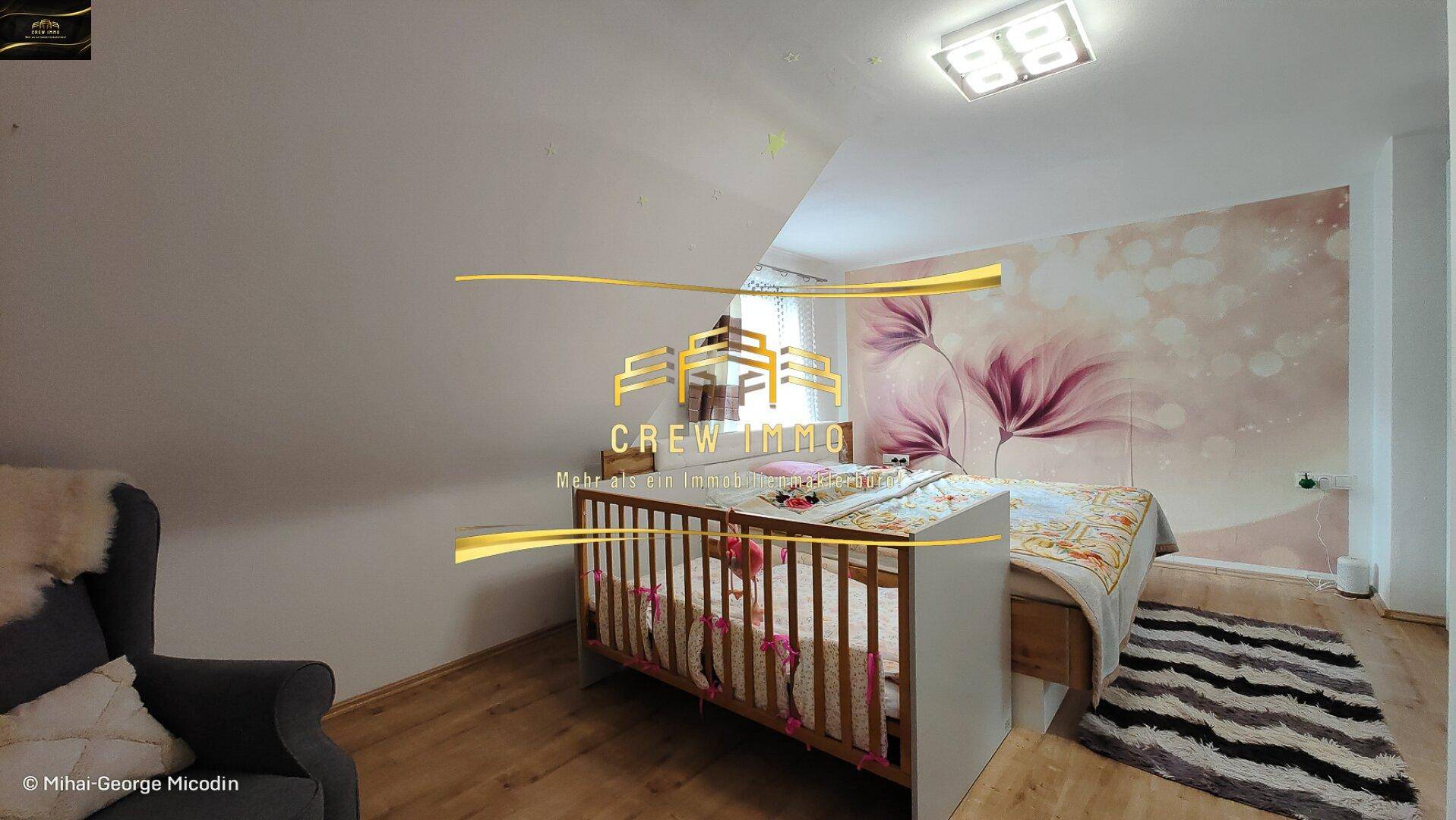 Helles Kinderzimmer im Dachgeschoss mit Babybett, schräger Decke und floraler Tapete.