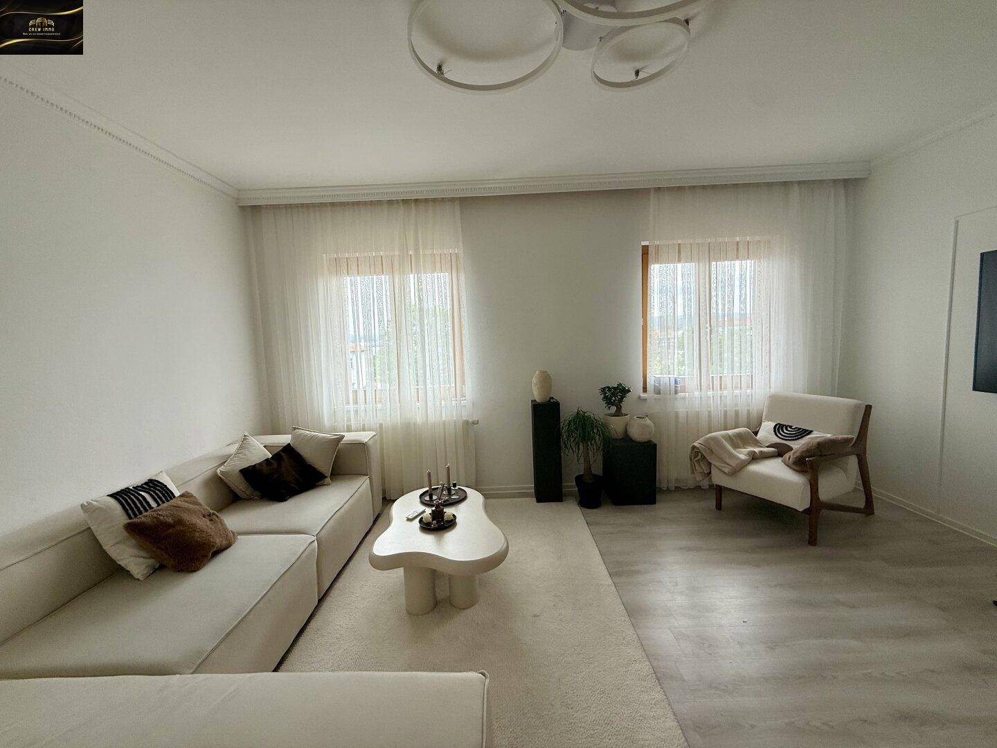 Geräumiges Wohnzimmer mit beigem Sofa, Sessel und zwei großen Fenstern für viel Tageslicht.