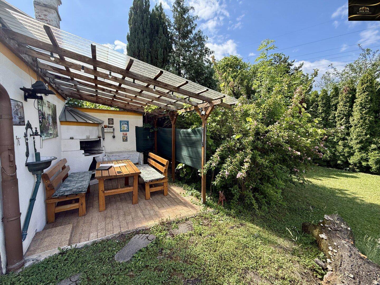 Überdachte Terrasse mit Holzbänken, Tisch und gemauertem Grillbereich im Garten.