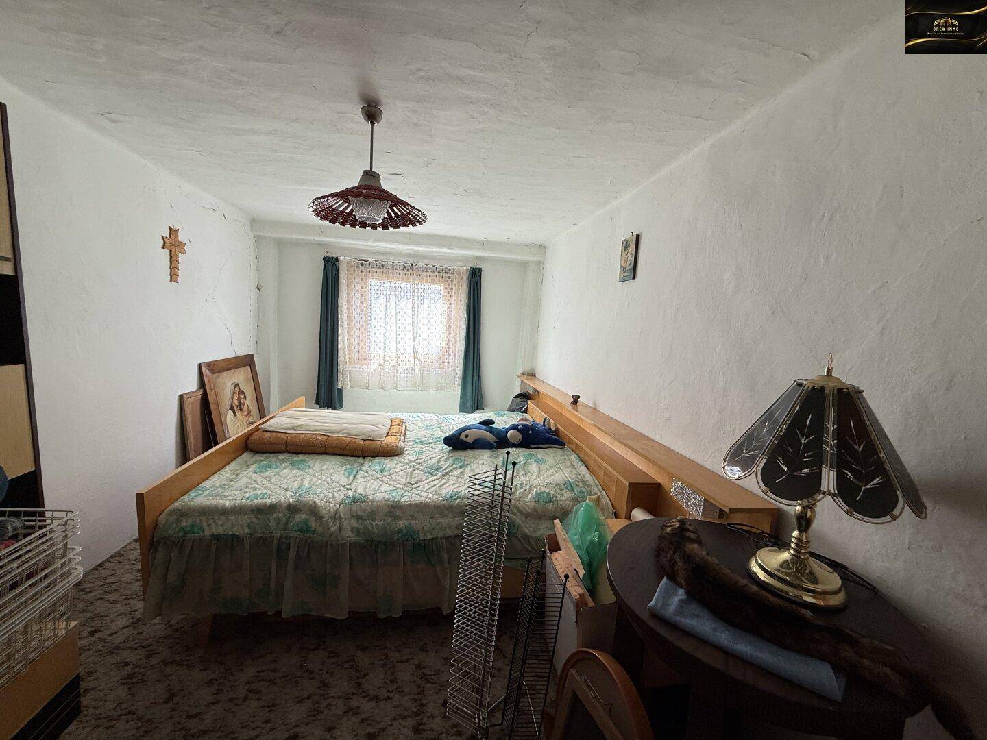 Schlafzimmer mit Bett, Fenster und einfacher Einrichtung, Wände mit rauer Putzstruktur.