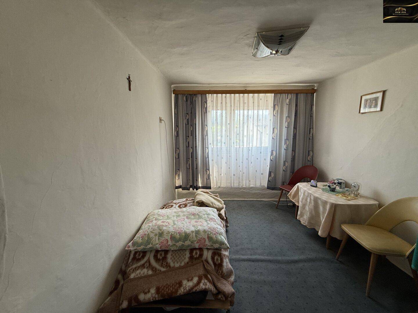 Schlafzimmer mit Bett, Fenster und einfacher Einrichtung, Wände mit rauer Putzstruktur.