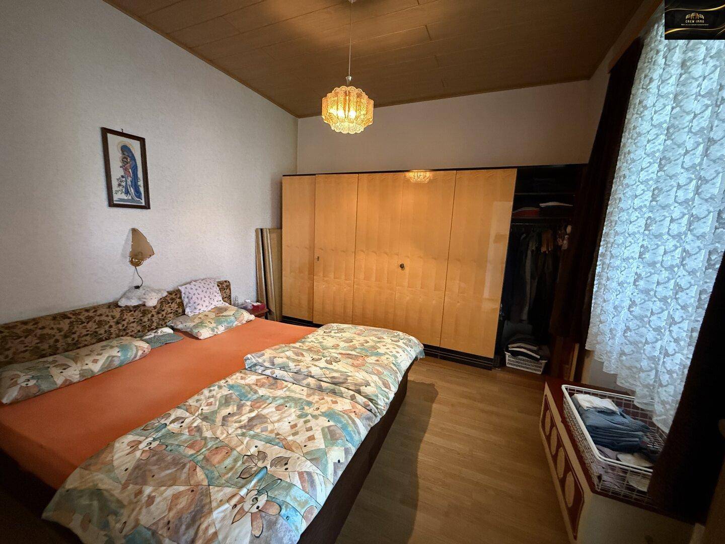 Schlafzimmer mit Bett, Holzschrank und Fenster, beleuchtet von einer Deckenlampe.