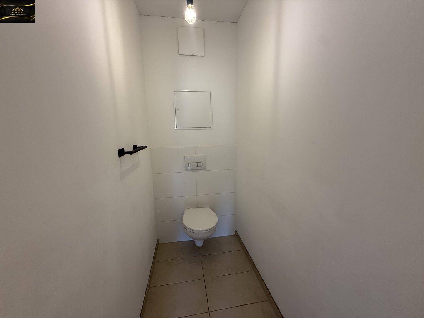 Separates WC mit Wand-WC, kleinem Waschbecken und Fliesenboden.
