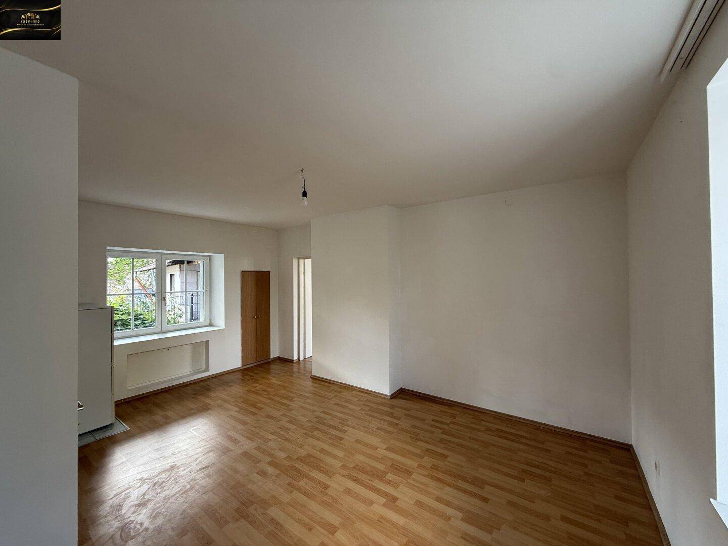 Großer, leerer Raum mit Laminatboden und einem Fenster, das den Blick nach draußen freigibt.