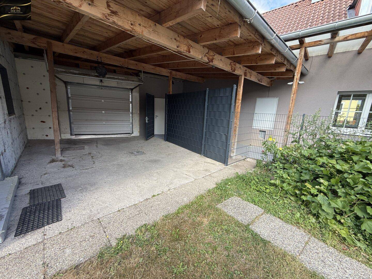 Überdachter Stellplatz neben einer Garage mit Holzbalken und einem kleinen Gartenbereich.