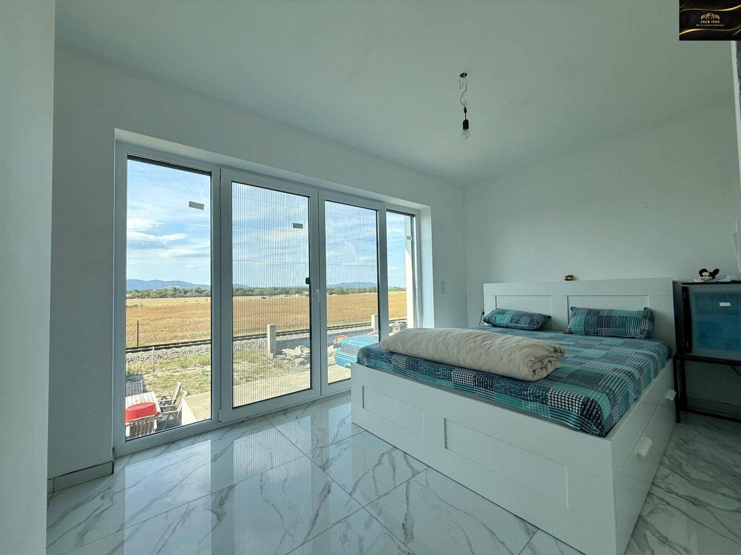 Helles Schlafzimmer mit großzügigen Fenstern, weißem Bett und direktem Zugang zum Außenbereich mit Weitblick.