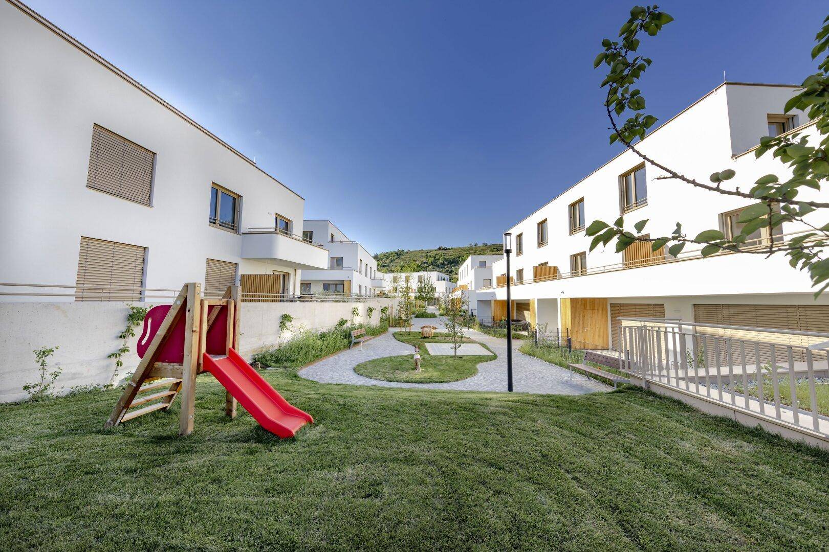 Moderne Wohnanlage mit weißer Fassade und einem Spielplatz mit roter Rutsche im Vordergrund.