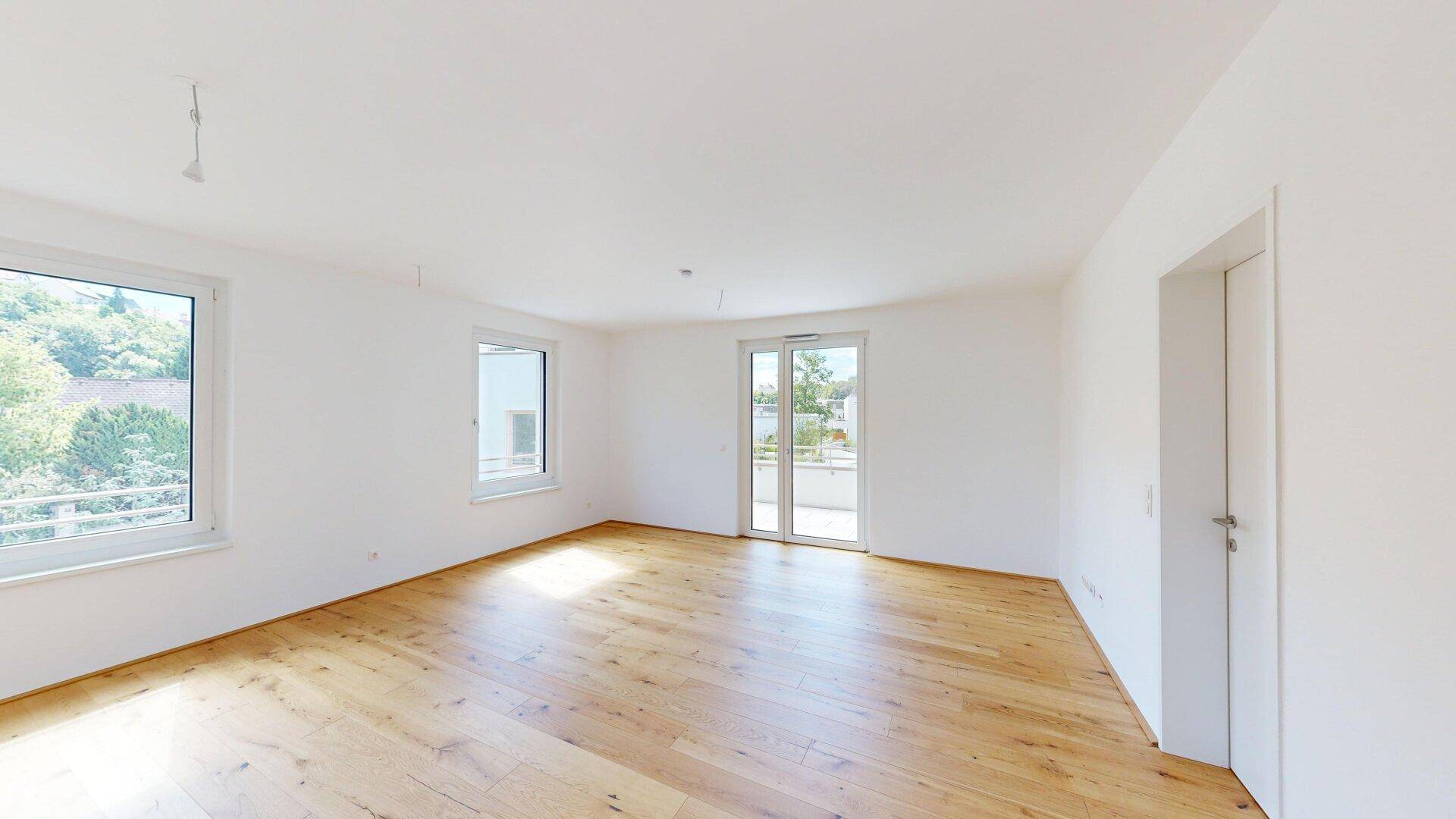 Heller, leerer Wohnraum mit Eichenparkettboden, großen Fenstern und Zugang zum Balkon.