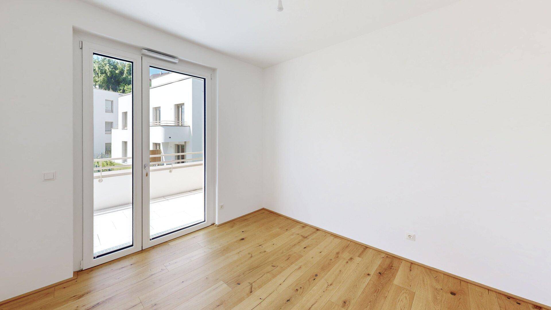 Unmöbliertes Schlafzimmer mit hellem Parkettboden und Balkontür, die viel Tageslicht hereinlässt.