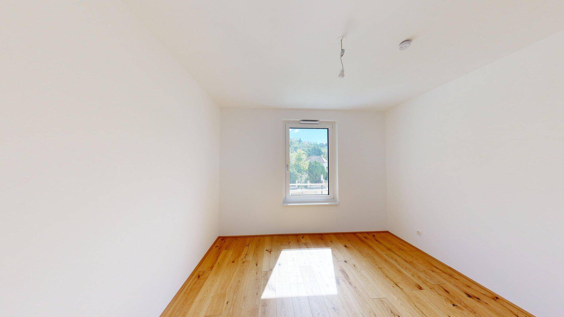 Schlichtes Schlafzimmer mit hellem Parkettboden, weißen Wänden und einem Fenster mit Ausblick.