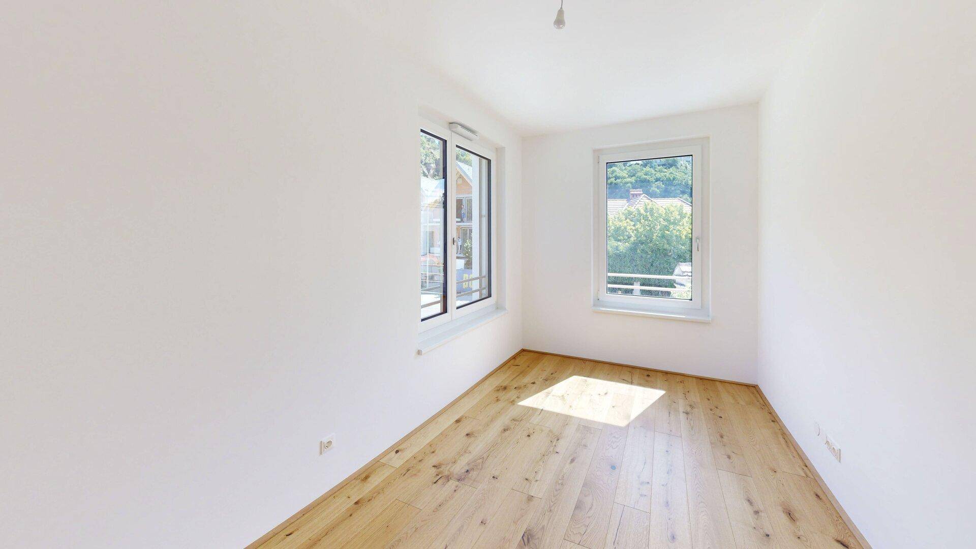 Helles Schlafzimmer mit zwei Fenstern, Parkettboden und weißen Wänden, ideal für viel Tageslicht.