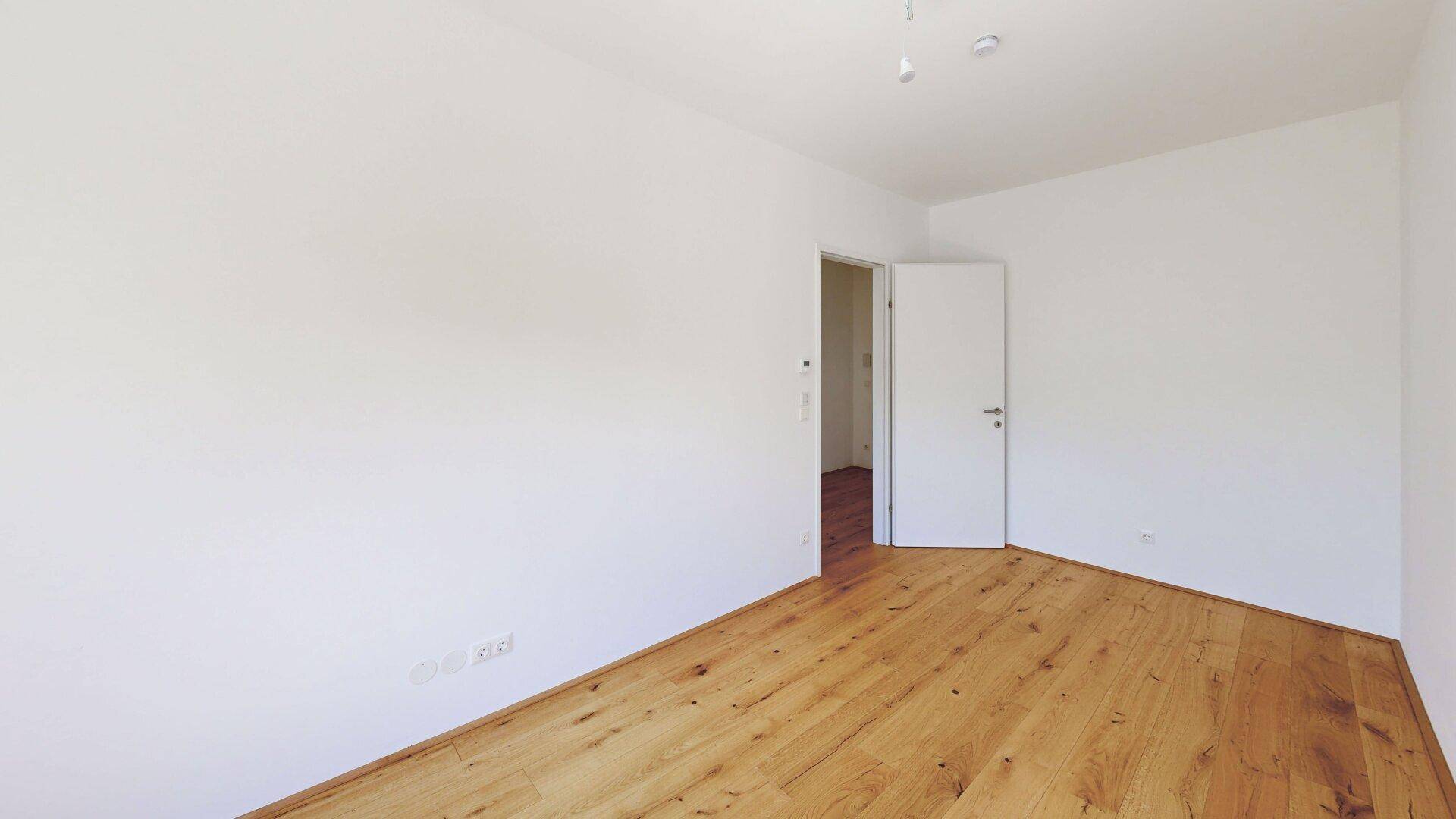 Unmöbliertes Zimmer mit Parkettboden und weiß gestrichenen Wänden, mit Zugang zu einem weiteren Raum.