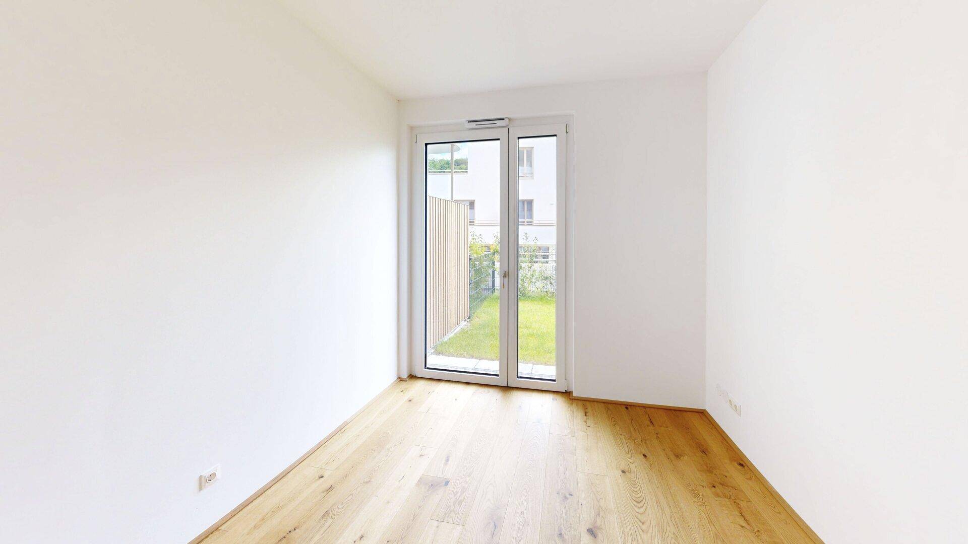 Leeres Zimmer mit Holzboden und großem Fenster, das einen Blick ins Grüne bietet.