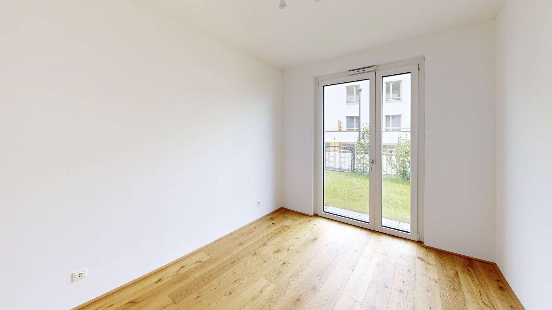 Helles, leeres Schlafzimmer mit Holzboden und Fensterfront zum Gartenbereich.