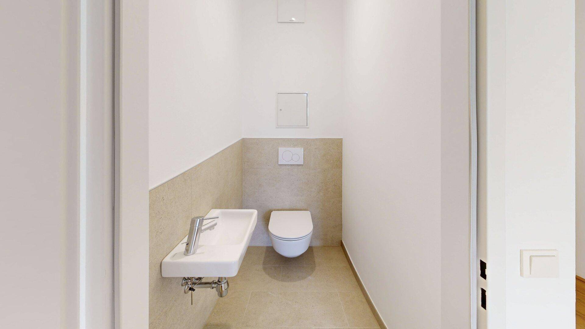 Separates WC mit wandhängender Toilette, kleinem Waschbecken und hellen Fliesen.
