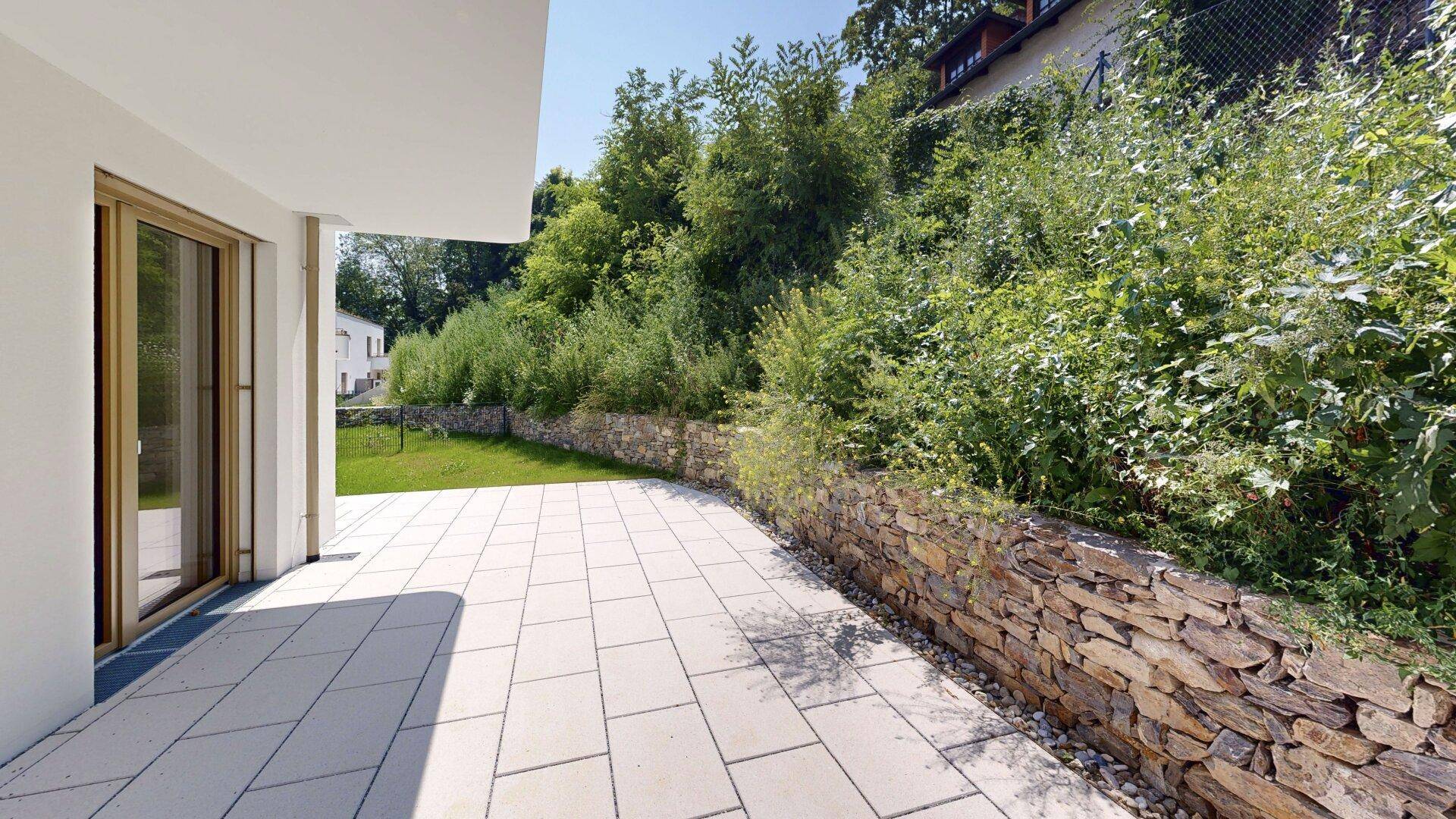 Große gepflasterte Terrasse mit Steinmauer und Blick auf grüne Hügel und Bäume.