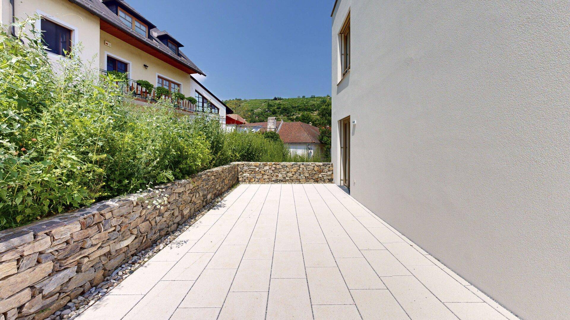Lange, gepflasterte Terrasse entlang des Gebäudes mit Steinmauer und Blick auf die Umgebung.