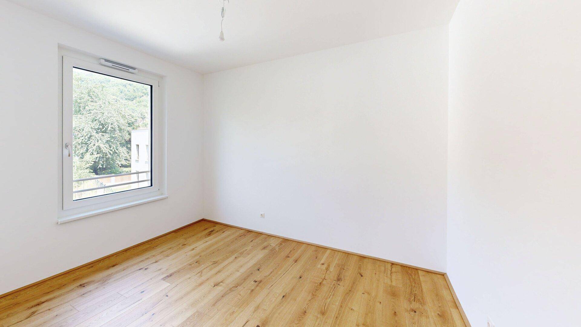 Helles, unmöbliertes Schlafzimmer mit Holzboden und einem Fenster mit Blick ins Grüne.